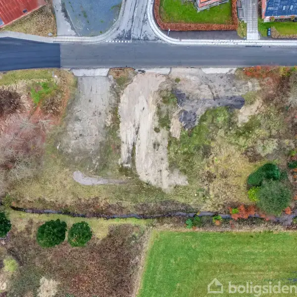 Grusbelagt grund ligger ved asfalteret vej, omkranset af græsarealer og huse. Vegetation ses langs kanten med grønne buske og træer.