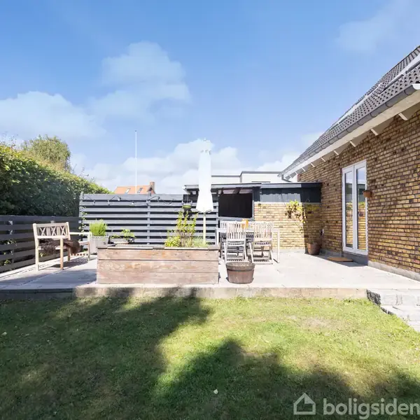 En terrasse med havemøbler og en parasol står ved siden af et hus med murstensvægge. Græsplæne og hæk omkranser terrassen. Der er ingen synlig tekst bortset fra "Boligsiden".