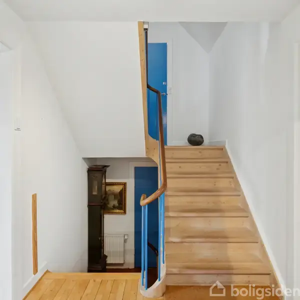 Trætrappe stiger op med et blåt gelænder i en lys entré. Stuen til venstre indeholder et bord og moderne kunst på væggen.