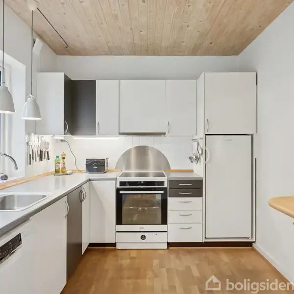 Et moderne køkken med hvide skabe og trælaminatgulve. En ovn, indbygget køleskab og vask sidder under vedhængslamper. Der står to flasker ved siden af vasken, en rund bordplade til højre.