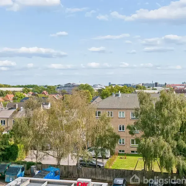 Udsigt over et byområde med lejlighedsbygninger og grønne træer. Handling ses ikke. Baggrund af byen strækker sig til horisonten under en blå himmel med spredte skyer.