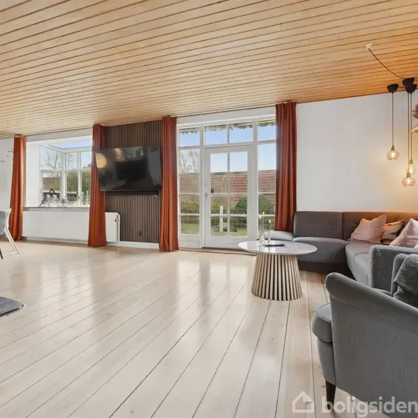 Stue med pejs opvarmer rummet; stor sofa og sofabord står i hjørnet. Vinduet med orange gardiner slipper lys ind. Flot trægulv og loftsspots fuldender den moderne indretning.