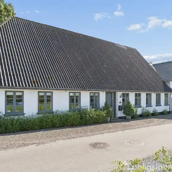 Hvid, langstrakt bygning med mørkt tag, har flere vinduer og en hoveddør. Omgivet af grønne planter, beliggende ved en grusvej under en klar blå himmel.
