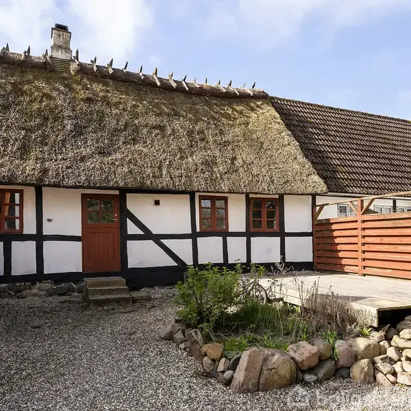 Bindingsværkshus med stråtag og træhegn står i en gårdhave omgivet af grusbelægning og blomsterbede. Huset har små vinduer og en enkelt trådør.