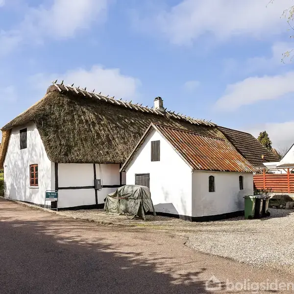 Et traditionelt bindesværkshus med stråtag står ved en vej. Huset er hvidt med en lille indhegning til højre. Omgivelserne er landlige med flere huse i baggrunden.