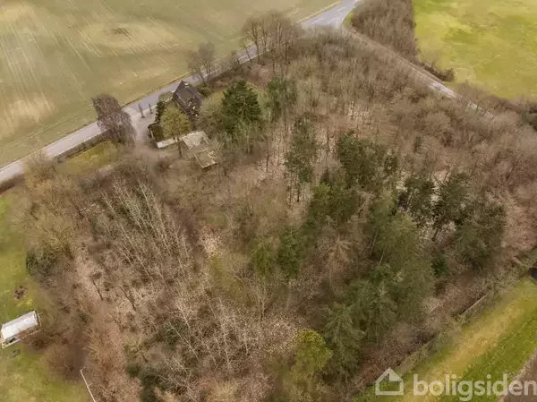 Skovklædt område omgiver en landejendom med bygninger. Veje ses omkring området, der ligger i en landlig kontekst med åbne marker. Træer dominerer landskabet nær huset.