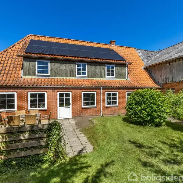 Rødt murstenshus med solpaneler på taget står i en frodig have. Foran huset er der en terrasse med havebord og stole. Til højre ses en ældre bygning.