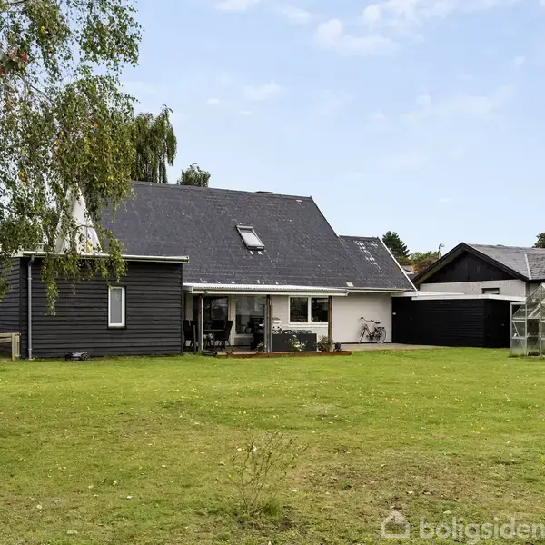 Hus med sort træfacade står stille omkranset af grøn græsplæne og et drivhus til højre. Himlen er blå, og en stor trækrone hænger ind i billedet fra venstre.