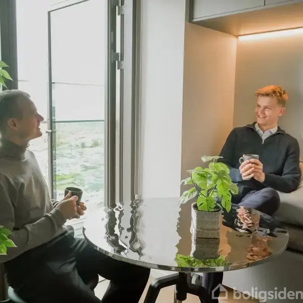 To personer sidder og taler, mens de holder kopper. De sidder i en moderne og lys lejlighed med store vinduer og grønne planter.