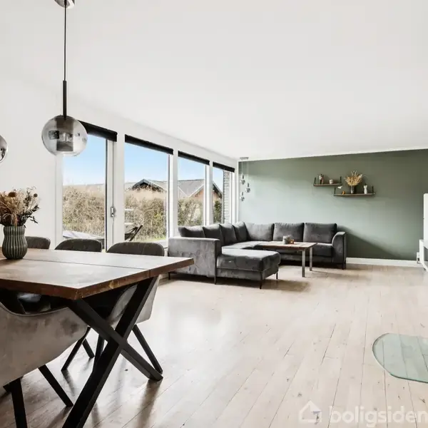 Spisebord med stoler står i en lys stue med trægulv. Store vinduer giver udsigt til udendørs haveområde. I baggrunden ses en grå sofa ved en grøn væg og en pejs.