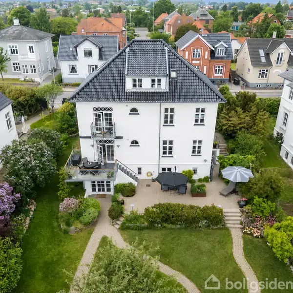 Hvidt, treetagers hus med skråt tag står i en velplejet have med stier og terrasse. Omkranset af lignende huse i et boligområde.