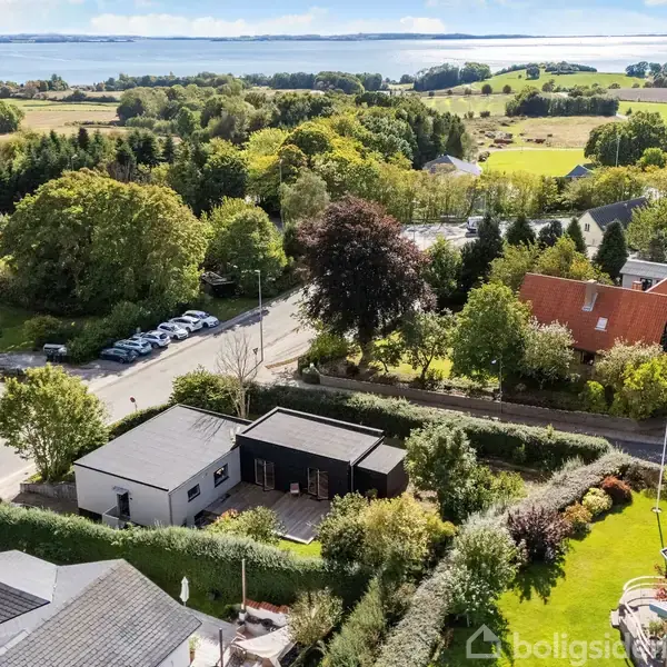 En moderne, sort og hvid bygning ligger blandt frodige haver og træer. Omgivet af huse og veje, med udsigt til et åbent landskab og vand i baggrunden.