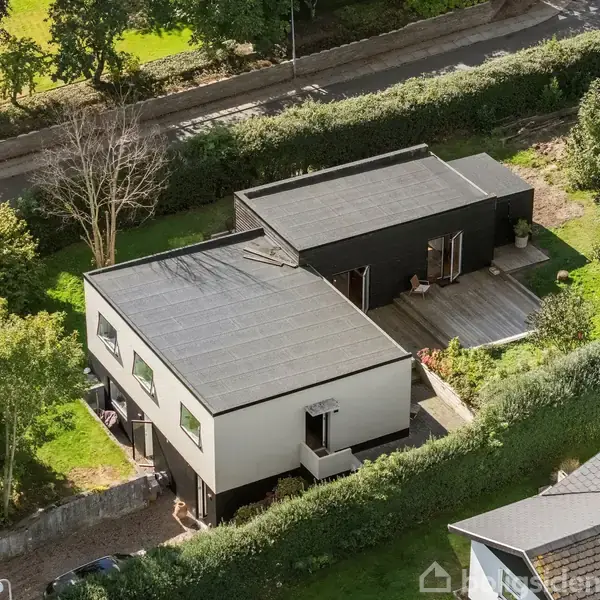 Et moderne hus set ovenfra ligger i en velplejet have omgivet af hække og træer. Huset har flade tage og store vinduer, med en terrasse i træ ved siden af.