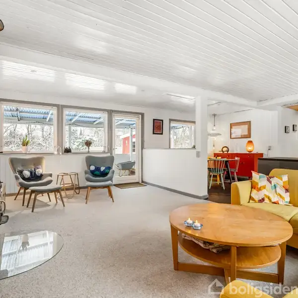 Brændeovn står stående, afgiver varme i et lyst, hyggeligt stueområde med gule sofaer, runde borde og store vinduer. Baggrunden inkluderer et åbent køkken og spiseplads.