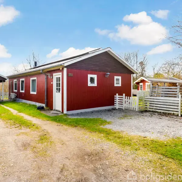 Et rødt hus med hvide detaljer står stille ved en grusvej. En carport til venstre og hegn omkredser haven i landlige omgivelser med blå himmel og spredte træer.