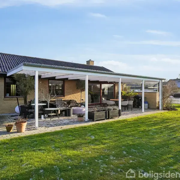 En overdækket terrasse med havemøbler står ved siden af et murstenshus. Græsplæne strækker sig ud foran, og der ses en hvid stakit i baggrunden under en blå himmel.