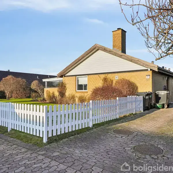 En gul murstenshus omringet af en hvid træhegn står stille i et villakvarter, med barne buske og en flisebelagt indkørsel op til huset.