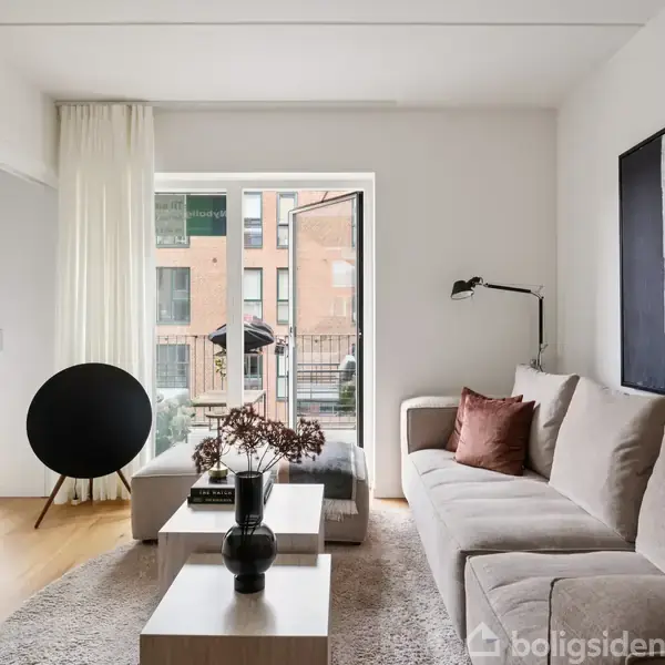 Beige sofa står i stue med hvidmalede vægge. Et sofabord med planter og lamper er i centrum. Et vindue med udsigt til murstensbygninger ses i baggrunden.