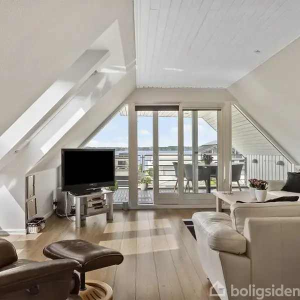 En lys stue med skrå loft og trægulv har en tv-skærm foran en lænestol. En sofa og et sofabord står mod en stor altandør med udsigt til en balkon.
