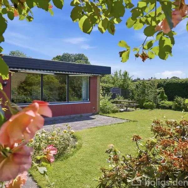 Hus med store vinduer står stille i en frodig have. Omkring er der græsplæne og farverige blomster under en klar blå himmel.