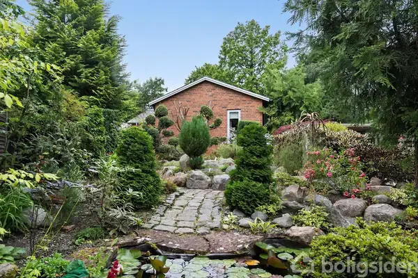 Et lille rødt murstenshus står i baggrunden af en frodig have. Foran huset ses en stensti omgivet af grønne buske og blomster, der leder til en dam med åkander.
