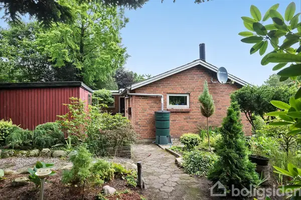 Rødt murstenshus med et fladt tag, omgivet af en frodig have med buske og træer. En smal stensti fører fra forgrunden til husets indgang.