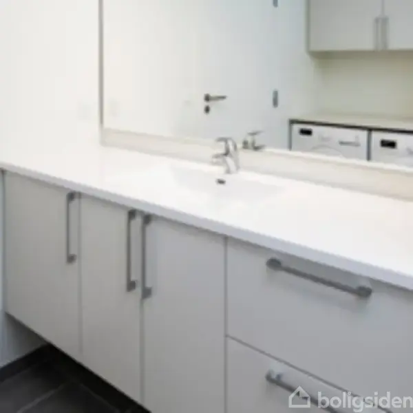 Håndvask med hvid bordplade, modernistiske skabslåger og spejl. Skabene har metalhåndtag. Et vaskeri set i spejlet, og rummet har flisegulv.