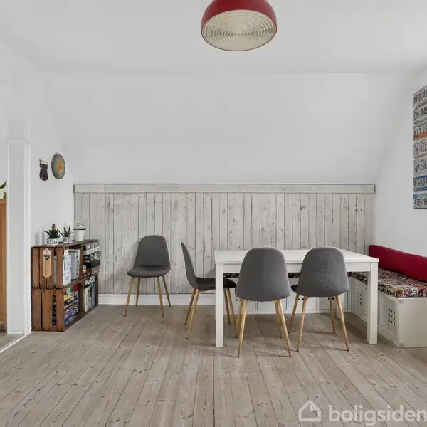 Et spisebord med grå stole står i et lyst rum med lyse trægulve. Et vindue til højre giver udsigt til omgivelserne. Væggene har hvidt panel og nogle farvede skilte.