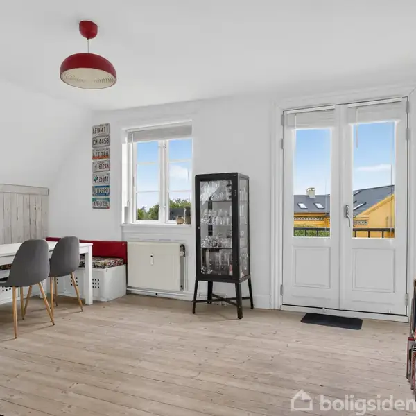 Et lyst rum med parketgulv. Et skrivebord med to stole står ved vinduet. En glasvitrineskab til højre. Dobbeltglasdøre fører ud til en lille balkon.