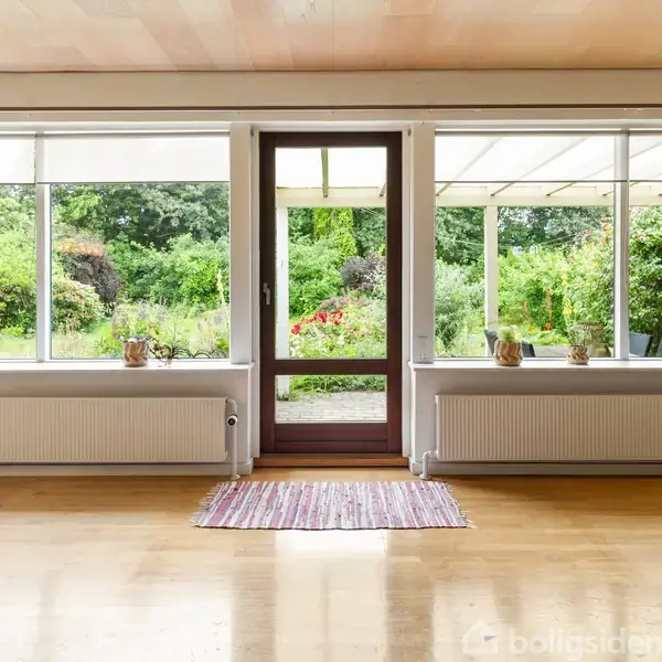 Dør med glas og to store vinduer giver udsigt til grøn have. Gardiner hænger ved siden af vinduerne. Trægulv og tæppe ses forrest. Ingen tekst findes.
