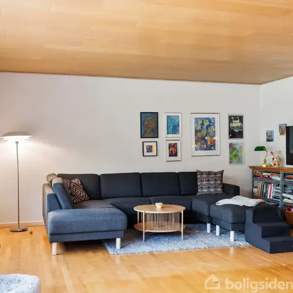 Sofaarrangement med sort hjørnesofa og rundt sofabord. Væggene prydes af kunst. Tv-reolen er fyldt med bøger. Store vinduer giver lysindfald. Trægulv og enkelt tæppe.