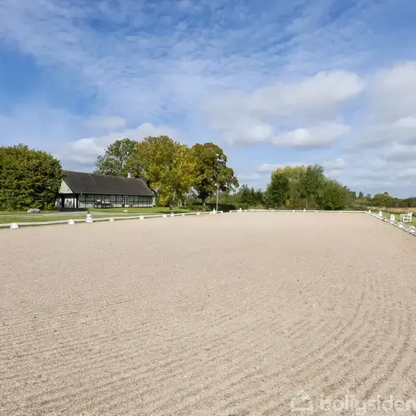 En stor, sandet ridebane er omgivet af grønt græs og træer under en klar blå himmel. I baggrunden ses et traditionelt sort bindingsværkshus.