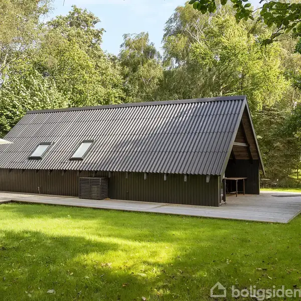 Et sort træhus med skråt tag står på en grøn græsplæne omgivet af træer. Ved siden af huset er en terrasse med parasol og havemøbler.
