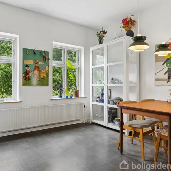 Spisebord med stole står i et lyst køkken. To lamper hænger over bordet, og der er en hvid reol ved siden af. Vinduet viser grøn udsigt. Plakater pryder væggene.