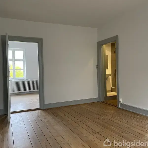 Et tomt rum med trægulv, to åbne døre; én fører til et andet værelse med et vindue og radiator, den anden til en gang.