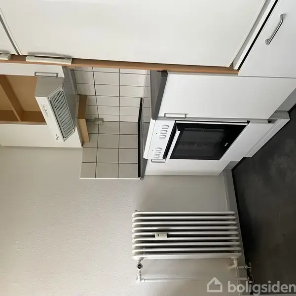 Hvidt køkken med komfur og ovn, flisevæg og hvide skabe. Til højre er der en grå bordplade. En radiator er synlig i forgrunden mod en lys væg.