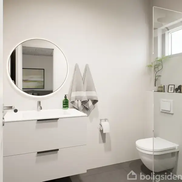 Et moderne badeværelse med et rundt spejl over en hvid vask. Ved siden hænger to håndklæder, og en grøn sæbepumpe står på vasken. Et toilet er placeret ved siden af.