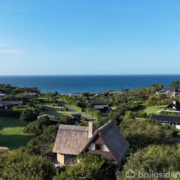Stråtækt hus ligger midt i en frodig grøn landsby med udsigt over havet i horisonten. Omgivet af buske, træer og flere huse langs veje.