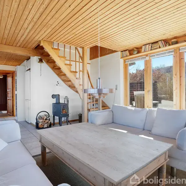 Stue med lyse sofaer omkring et stort træbord, pejs til venstre. Træloft og trappe fører opad. Store vinduer giver udsigt til haveområdet. Hylder over vinduerne.