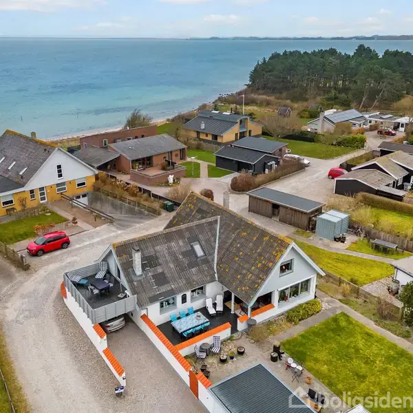 To-etagers hus med hvid facade og sort tag, omkranset af terrasse og grønt område, ligger tæt på kystlinje med udsigt over havet. Flere huse og en grusvej ses i nærheden.