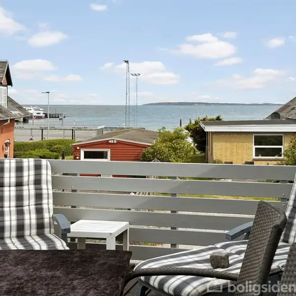 To terrassestole med sort-hvidt ternet mønster står på en altan, der vender mod havet. Omkringliggende bygninger rummer røde og gule facader.