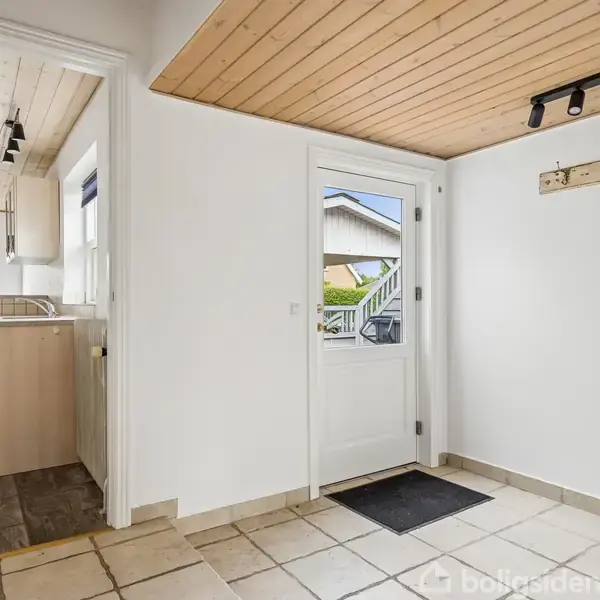 Dør står i en entre, som har flisegulv og en lys træloft. Hængende loftlampe og knagerække er installeret, mens en åben dør fører til køkkenet med træskabe.