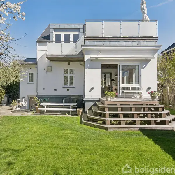 Hus med hvid facade og terrasse, omgivet af en velplejet græsplæne og træer. Solrigt vejr bidrager til en indbydende atmosfære i baghaven. Terrasse har trapper og siddepladser.