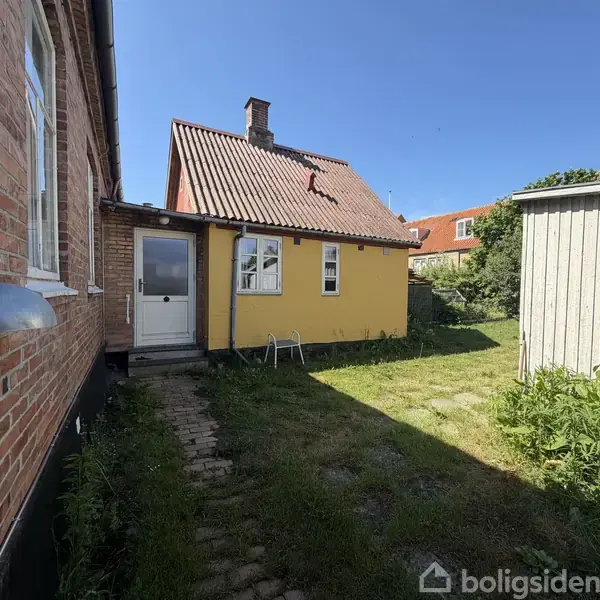 Gul murstensbygning med rødt tag står ved siden af en rød murstensbygning. Foran ses en have med græs og enkelte buske. Vejret er solrigt med blå himmel.