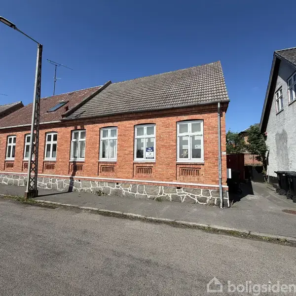 En rødstensbygning med flere vinduer står langs en stille gade. Bygningen har et skråt tag og en lys facade. En lygtepæl står på fortovet foran huset.