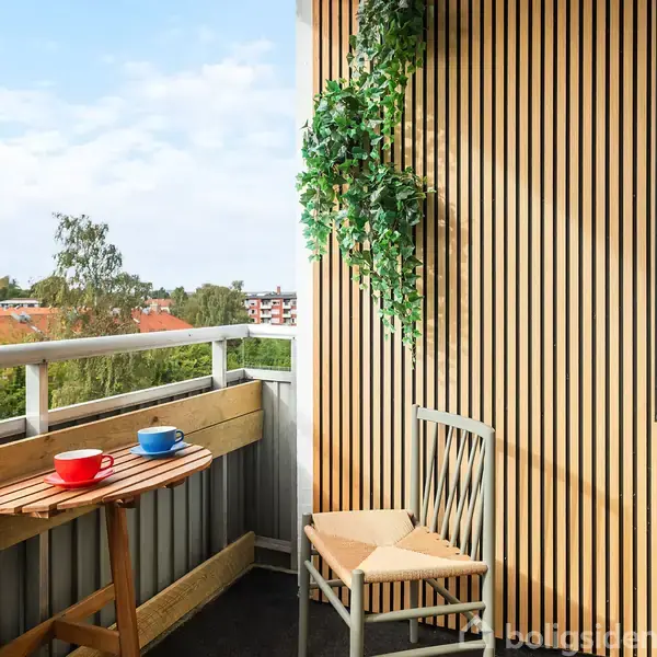 En lille balkon med et træbord, hvorpå der står en rød og en blå kop. En træstol står ved siden af, omgivet af en planteranke på væggen, med udsigt over trætoppe og hustage.