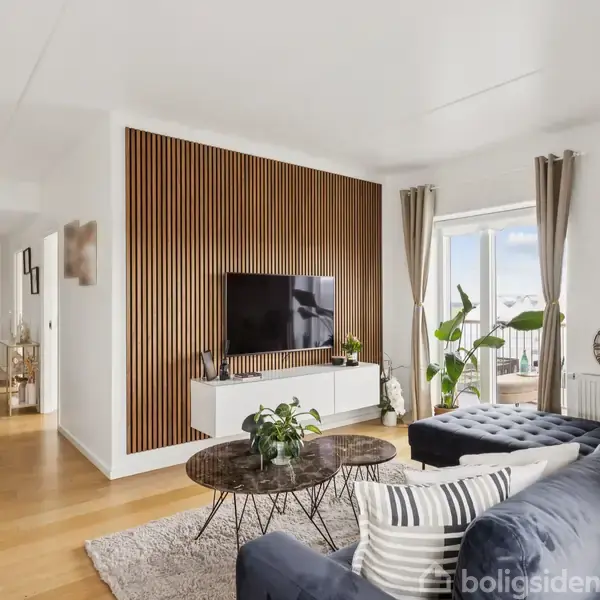En stue med en blå sofa og sofabord foran et vægmonteret TV på en træbeklædt væg. Store vinduer med gardiner viser udsigt til altanen.