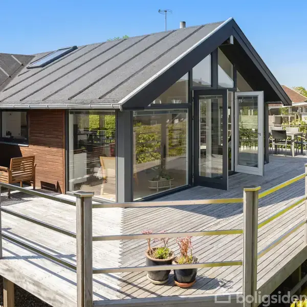 Et moderne sommerhus med store vinduer og en skrånende tagkonstruktion. En træterrasse omgiver huset med siddemøbler og planter i potter. Baggrunden har grønne hække og en lys, blå himmel.