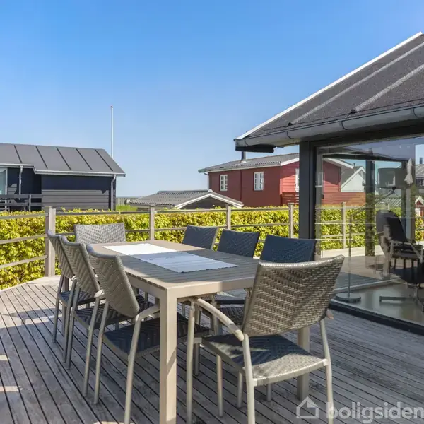 Udendørs terrasse med spisebord og stole i flettet design. Bordet står ved siden af et moderne hus med store vinduer. En hæk omgiver terrassen under en klar blå himmel.