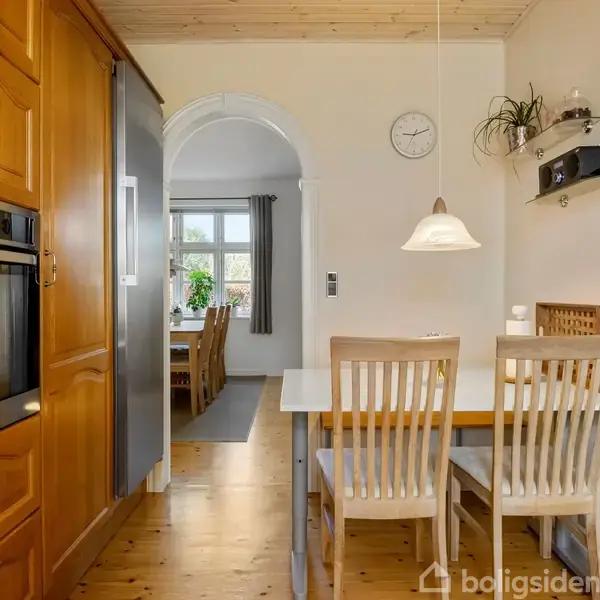 Et køkken med træskabe har en bordplade med to stole ved et bord. I baggrunden ses en spisestue gennem en buegang. Vægur og hylder hænger på væggen.